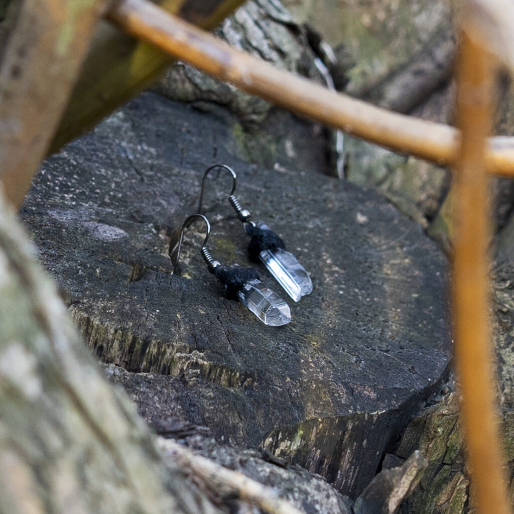 Nahaufnahme von kleinen Ohrhängern mit klaren Bergkristallspitzen und schwarzem Hanfgarn in rustikaler Umgebung auf einem Baumstumpf.