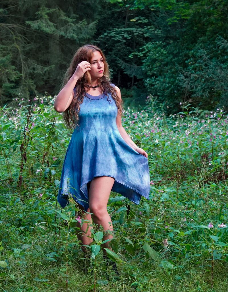 Blaues handgefärbtes Baumwollkleid mit weitem Zipfelrock, getragen von einer Frau mit wildem Haar auf einer Lichtung im Wald, umringt von Wildblumen. Das Kleid hat eine A-Silhouette.