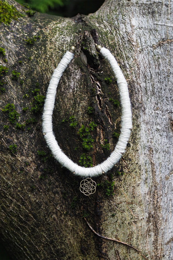 Dickes weißes Halsband aus Hanfgarn mit Metallperlen und geometrischen Anhänger, präsentiert an einem mit Moos bewachsenen Baum. Der Anhänger stellt den Samen des Lebens aus der Heiligen Geometrie dar.