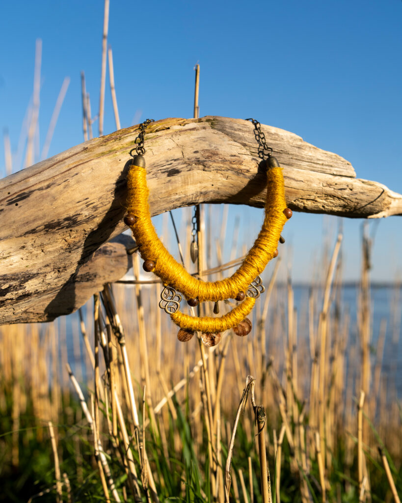Collier aus senfgelbem Hanfgarn mit Details aus Holz, Muschel und Altmessing, präsentiert auf einem Treibhaus zwischen kahlen Schilfhalmen vor einem Gewässer.