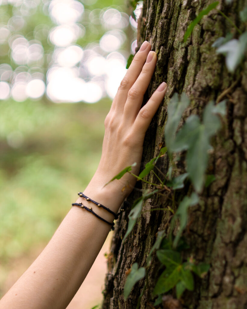 Zwei dünne schwarze Armbänder aus Hanf mit Altmessing-Perlen, getragen an am Handgelenk einer Hand, die an einem mit Efeu bewachsenen Baumstamm ruht.